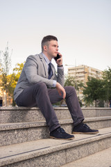 one young Caucasian man, blank expression, business suit, formal wear, ordinary common person portrait, outdoors sitting on stairs steps, talking listening, smart phone, ear