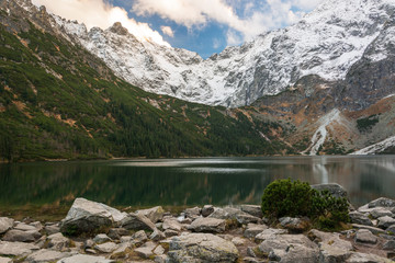 The landscape of  lake surrounded by snow mountains with  green fir-trees  and  blue sky with white clouds and big stones in the foreground