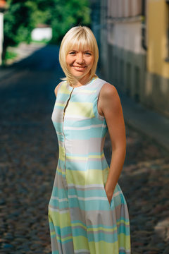Young stylish pretty blonde woman in beautiful colourful dress standing on the street in old town