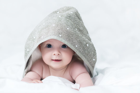 Cute baby girl or boy after shower with towel on head in white sunny bedroom. Child with big blue eyes smiling and relaxing in bed after bath or shower