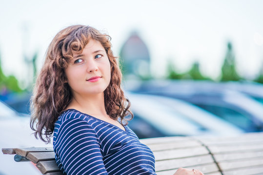 Portrait Of Young Brunette Woman Looking Over Shoulder While Sitting On Bench In Outdoor Park