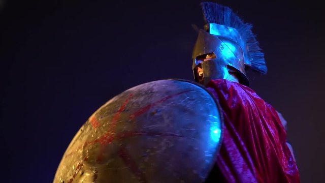 Roman gladiator in leather armor, helmet and red raincoat is covered with a round shield and strikes with a sword, slow motion shooting