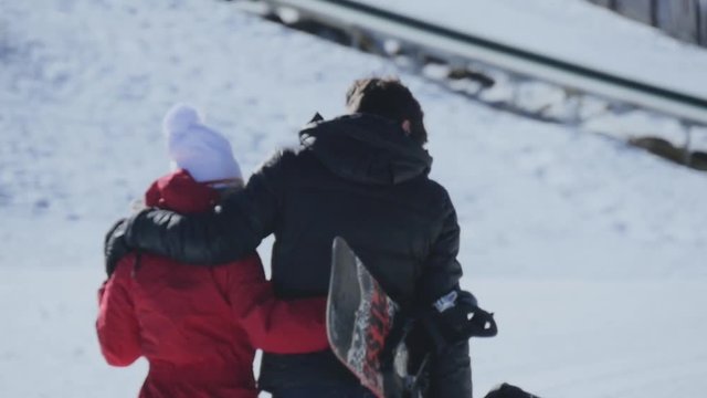 Girl walking hugging boy with snowboard on snow, in sky station