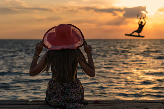 Beautiful girl in a red hat smiling at the summer waterfront and watching on the man kitesurfer. Silhouetted photo in the sunlights at sunset.