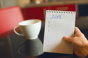 Notepad in hand with a plan for 2018, against a background of mugs with coffee