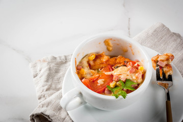 Trendy modern food, fast food, lunch in the microwave. Pizza in a mug, with ham, tomatoes, mushrooms, cheese. White cup, with a fork, on a white marble table. Copy space