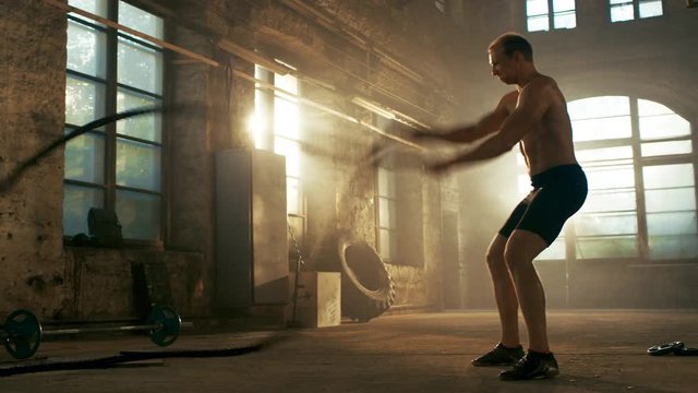 Strong Muscular Shirtless Sportsman Works out Hard with Battle Ropes as Part of His Cross Fitness Training Routine. He's Covered in Sweat and Exercises in a Abandoned Factory Remodeled into Gym.