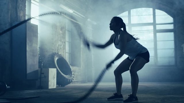 Strong Athletic Woman Exercises with Battle Ropes as Part of Her Cross Fitness Gym Workout Routine. She's Covered in Sweat and Training Takes Place in a Abandoned Factory Remodeled into Gym. 