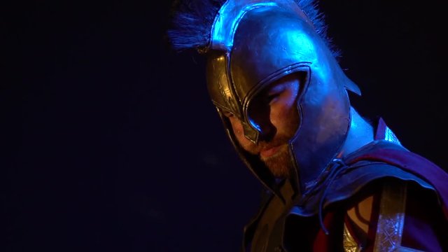 The sweaty Roman gladiator in leather armor, helmet and red raincoat stands with a pensive look and then turns to the camera