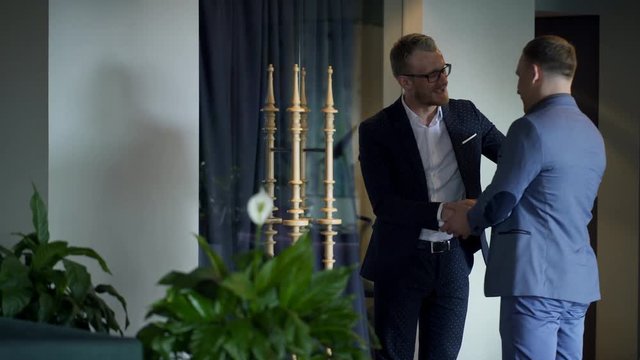 Young man stands and meets his colleague in hall of restaurant. Businessmen handshake each other and go to the table at the business lunch. They begin to talk after greeting. Handsome partners are
