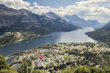 Naklejka na ściany i meble horizontal image taken from a mountain top looking down far below on to a small town of Waterton with a lake surrounded by mountains at Waterton National Park in the summer.