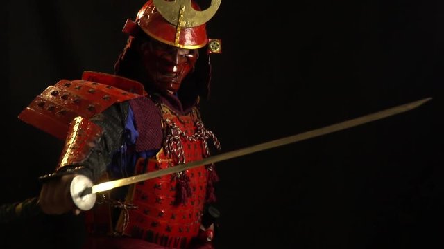 A samurai man in a beautiful red armor, helmet and red defensive mask of a demon pulls out a sword from its scabbards on a dark background, a bit slow motion