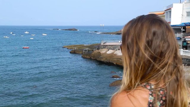 Tenerife. Spain. Beautiful blond girl on vacation observes ocean and restaurants in La Caleta village, Canary Islands. Girl Travels. .