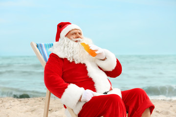 Fototapeta premium Authentic Santa Claus with bottle of drink relaxing in deck chair on beach