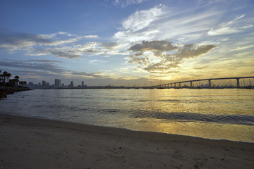 sand and palm trees line the shore of Coronado Island as the sun slowly rises above the Coronado Bridge