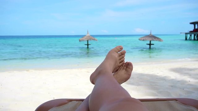Tourist legs bare feet sitting on golden sand beach bench point out to tropical ocean paradise