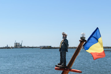 A sailor toy and the blue, yellow and red romanian flag mounted on a ship's mast. The Black Sea in the background