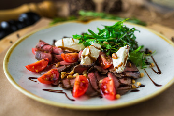 On a plate, on a wooden surface, close-up roast beef salad with rocket salad, cherry tomatoes, mozzarella, balsamic sauce, rosemary, spices, nuts, tall kitchen, restaurant, trendy delicious food
