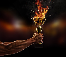 Muscular man arm holding burning trophy cup on black background