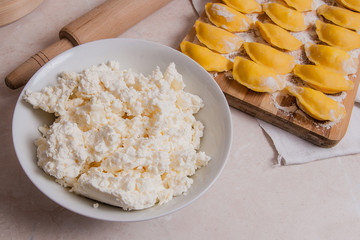 Ready for boiling vareniki, dumplings, pierogi on wooden cutting board with flour and white plate with cottage cheese or curd..