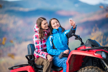 Fototapeta na wymiar Close-up two happy girls in winter jackets on red ATV smiling and makes selfie on the phone with blurred background nature