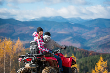 Guy driving ATV, girl sitting behind him and turned around, looking at the camera and smiling. Beautiful landscape mountains at autumn sunny day on blurred background