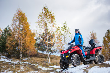 Happy woman in blue winter clothes riding on a red quadbike atv on snow-covered hill against autumn nature on the background with copy space