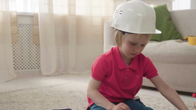 small blonde kid boy imitates engineer constructing a building using colorful meccano details with enthusiasm and focus dreaming about future profession, sitting at home on floor