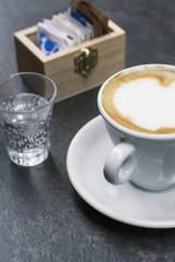 White cup of cappuccino with small glass of water on the grey table