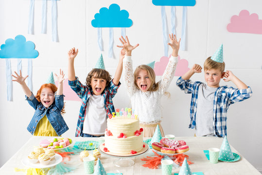 adorable happy kids raising hands and smiling at camera at birthday table