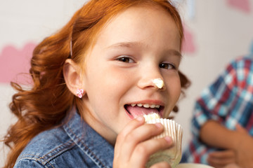 adorable red haired girl eating delicious cupcake and smiling at camera