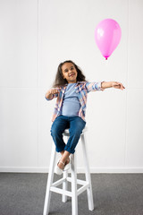 cute smiling african american girl holding balloon while sitting on stool at birthday party