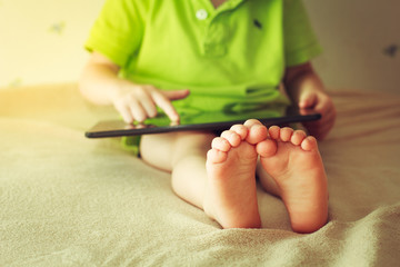 Kid with tablet sitting on bed and watching cartoons. Focus on the feet