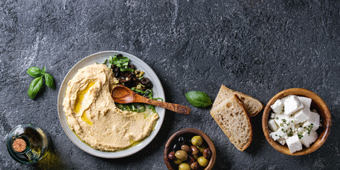 Homemade traditional spread hummus with chopping olives and herbs on blue plate, served with bread, olives, feta cheese, olive oil, spoon on black texture background. Mediterranean snack. Flat lay