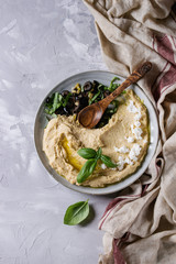 Homemade traditional spread hummus with chopping olives, oil and herbs on blue plate, served with feta cheese, spoon, basil, textile on gray texture background. Mediterranean snack. Flat lay