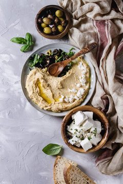 Homemade traditional spread hummus with chopping olives, oil and herbs on blue plate, served with bread, olives, feta cheese, spoon, textile on gray texture background. Mediterranean snack. Flat lay