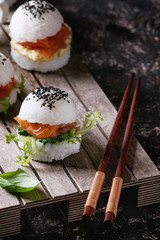 Mini rice sushi burgers with smoked salmon, green salad and sauces, black sesame served on wood pallet tray with chopsticks over dark brown concrete background. Modern healthy food. Close up