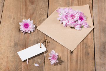 Blank white paper tag with brown envelope and pink flowers on wooden table.