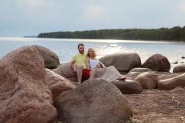 A man in shorts and a t-shirt and a girl in a summer dress enjoying each other. Beautiful couple sitting on a rock on the beach, watching the sunset. They wear casual clothes