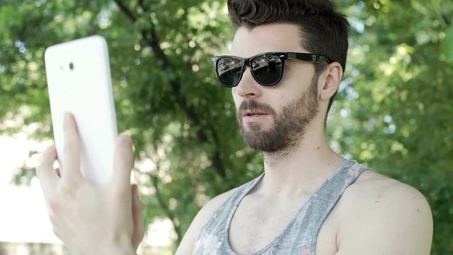 Handsome man in camis shrit standing in the park and doing selfies on tablet, steadycam shot
