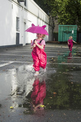 Children run through puddles on the street after a rain with pink umbrellas in hands, in pink bright rubber boots. The rays of the sun are reflected in the drops of water