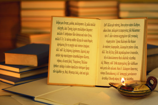 Open Book Illuminated By An Oil Lamp. The Opening Lines Of The Odyssey Written In The Ancient Greek Language. A Stack Of Books. On The Lamp, Visible Inscription Aphrodite In The Greek Alphabet.