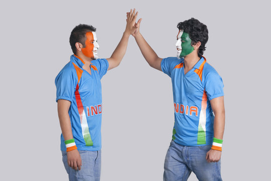Male cricket fans with face painted in Indian tricolor giving a high-five to each other over colored background 