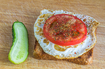Healthy breakfast: fried egg on a whole wheat toast with a slice of tomato, seasoning and fresh green cucumber on a wooden chopping board