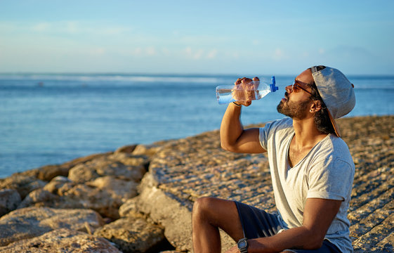 Young African Man Drinking Water On The Sea Beach.