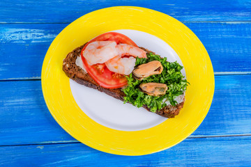 Sandwich with tomato, shrimps, mussels and chopped parsley on a yellow and white plate on a blue wooden background.