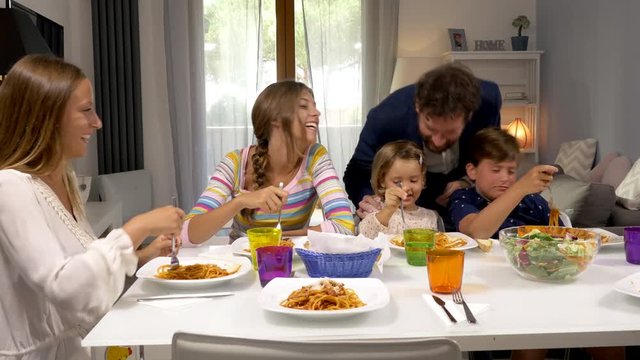 Father coming from work at home kissing kids and teenage daughters sitting on table eating spaghetti