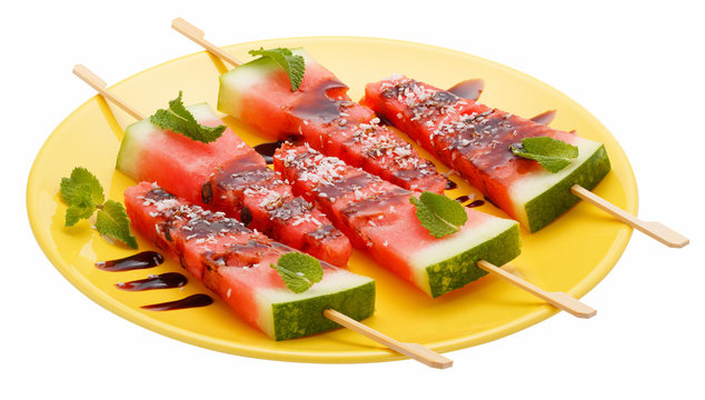Slices of watermelon with stick on yellow plate isolated on white background. It looks like ice cream. Decorated with mint leaves, coconut and chocolate syrup.