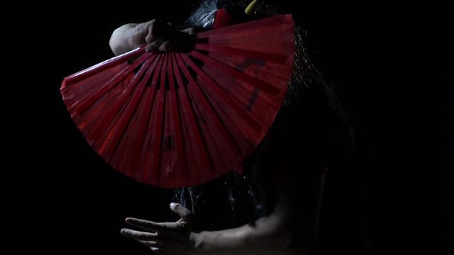 The freak in the mask of the Japanese demon and dressed in a black kimono covers his face with a red fan popping out of the darkness
