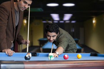 Young man playing snooker while friend standing besides 